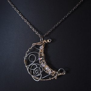 A crescent moon pendant crafted from silver wire with light gold details gracefully hangs from a delicate silver chain.