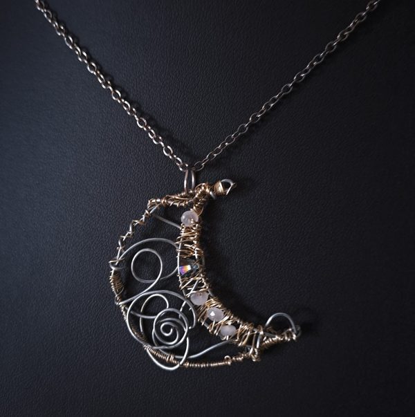 A crescent moon pendant crafted from silver wire with light gold details gracefully hangs from a delicate silver chain.