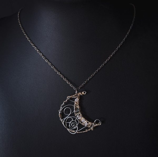 A crescent moon pendant crafted from silver wire with light gold details gracefully hangs from a delicate silver chain.