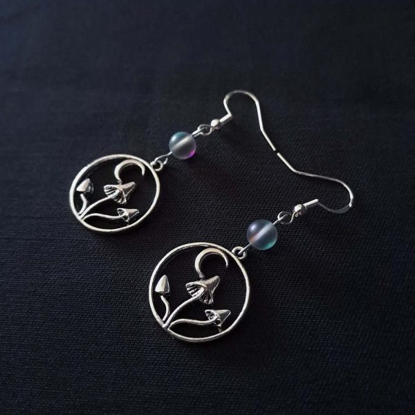 Nocturnal mushroom earrings. A stainless steel charm contains three rising mushrooms, and a half moon. Above the charms lays a translucent iridescent grey-blue bead, reminiscent of moonstone. Very witchy and gothic vibes