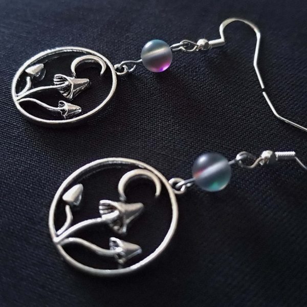 Nocturnal mushroom earrings. A stainless steel charm contains three rising mushrooms, and a half moon. Above the charms lays a translucent iridescent grey-blue bead, reminiscent of moonstone. Very witchy and gothic vibes