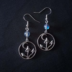 Nocturnal mushroom earrings. A stainless steel charm contains three rising mushrooms, and a half moon. Above the charms lays a translucent iridescent grey-blue bead, reminiscent of moonstone. Very witchy and gothic vibes