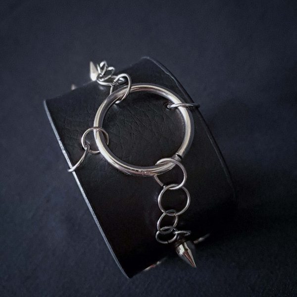 Black leather wristband with stainless steel spikes in assymetrical placement. Intertwined rings forming a chain and connecting th center large steel o-ring. A unique twist to a classis alternative fashion accessory of gothic or punk styles.